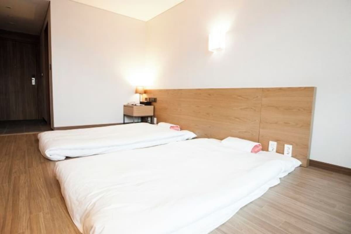 A hotel room with two beds, a wooden headboard, a nightstand with a lamp, and a wall-mounted light fixture.