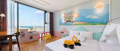 A hotel room with a large bed, a desk with a chair, a kitchenette, and a view of the ocean.