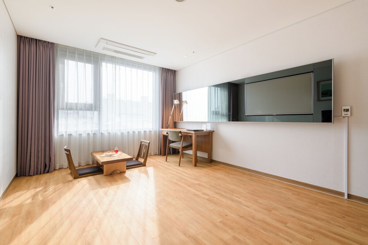Habitación de hotel moderna con suelo de madera, un escritorio con silla, un televisor de pantalla plana en la pared y un ventanal con cortinas.