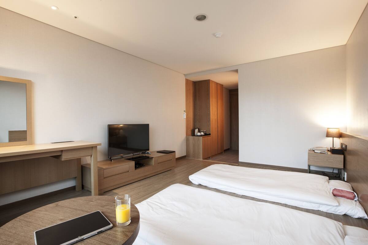 Hotelkamer met bureau, televisie, twee bedden, nachtkastje met lamp en een klein tafeltje met een glas sap en een tablet.
