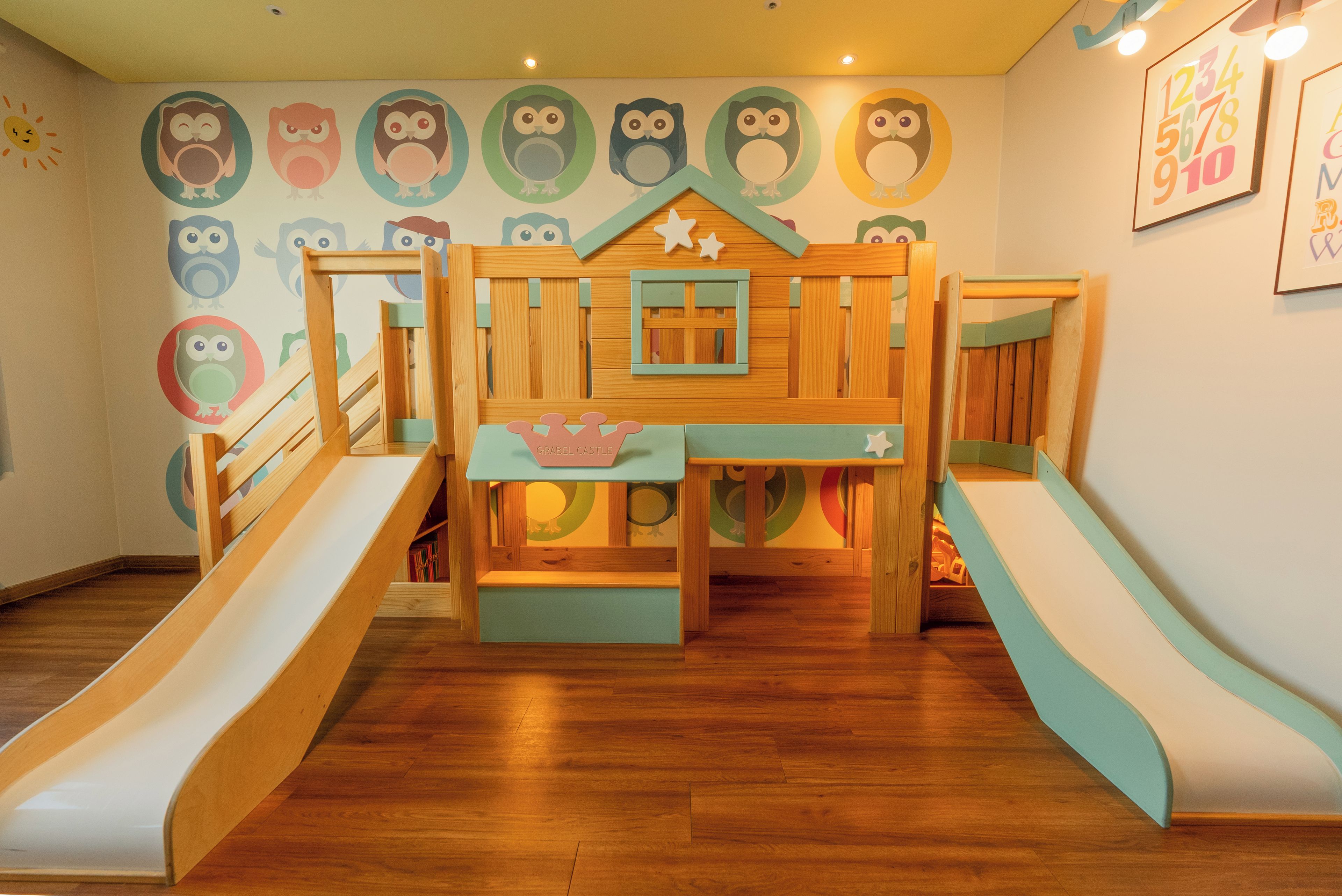 Un espace de jeux pour enfants avec un toboggan, une structure d’escalade et un mur décoré d’illustrations de hiboux.