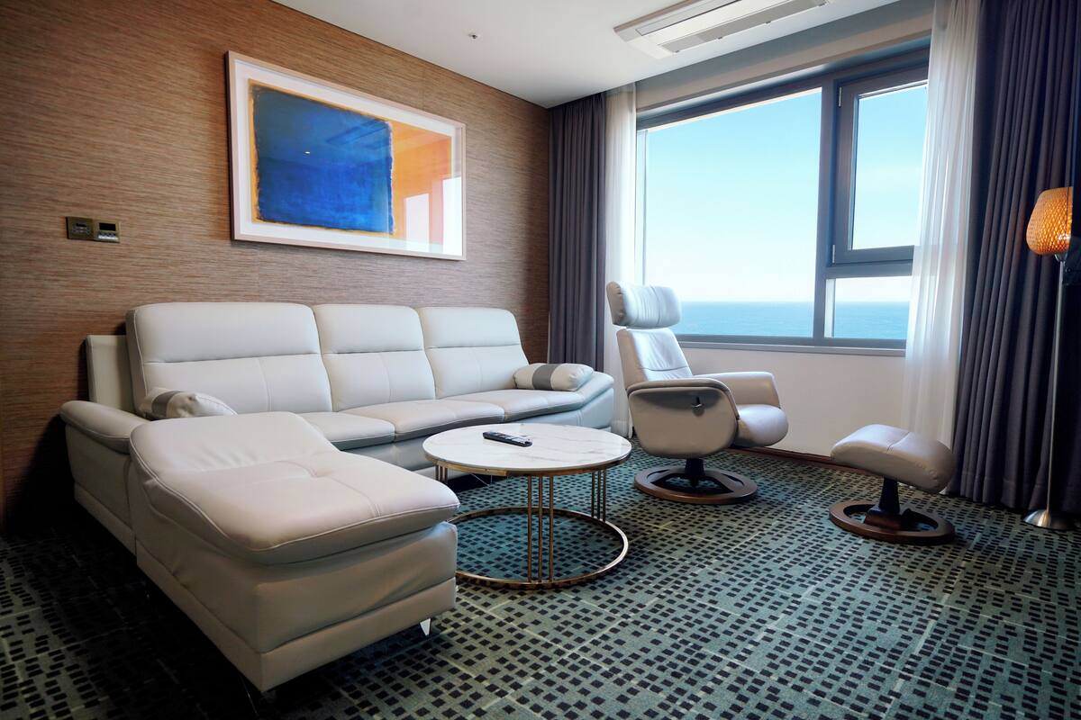 Een moderne hotelkamer met een bank, twee fauteuils, een klein bijzettafeltje en een groot raam met uitzicht op zee.