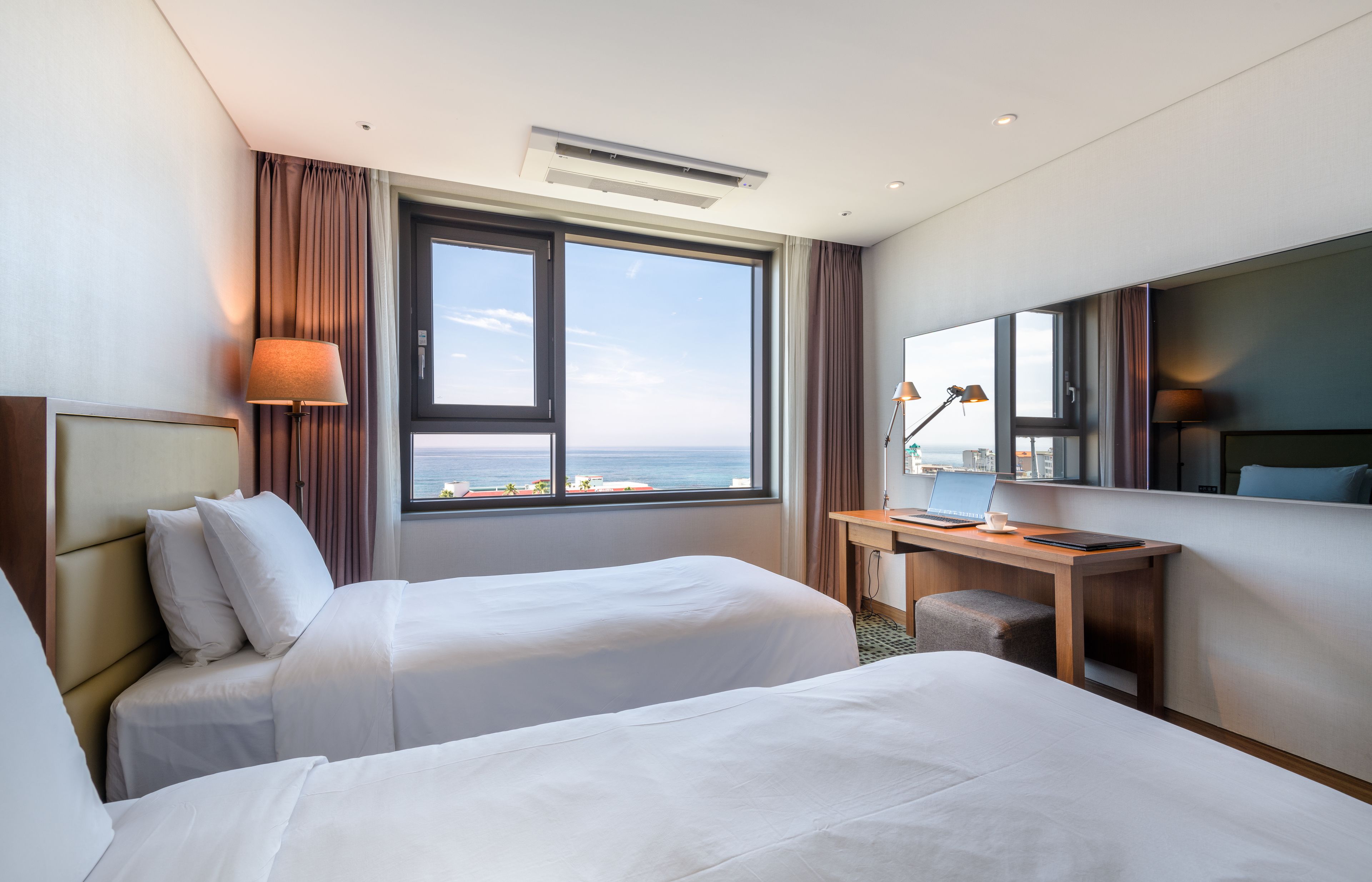 Habitación de hotel con dos camas, un escritorio, una silla, un ventanal con vista al mar y aire acondicionado en el techo.