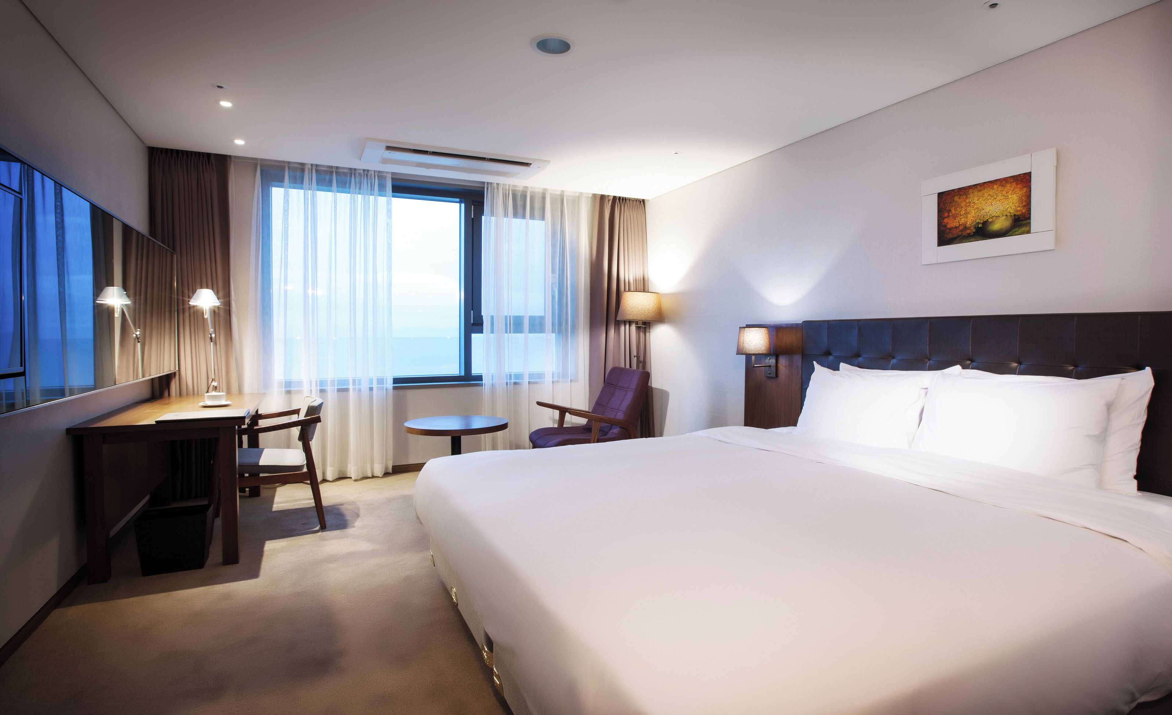 A hotel room with a large bed, a desk with a chair, a small table, and a window with curtains.