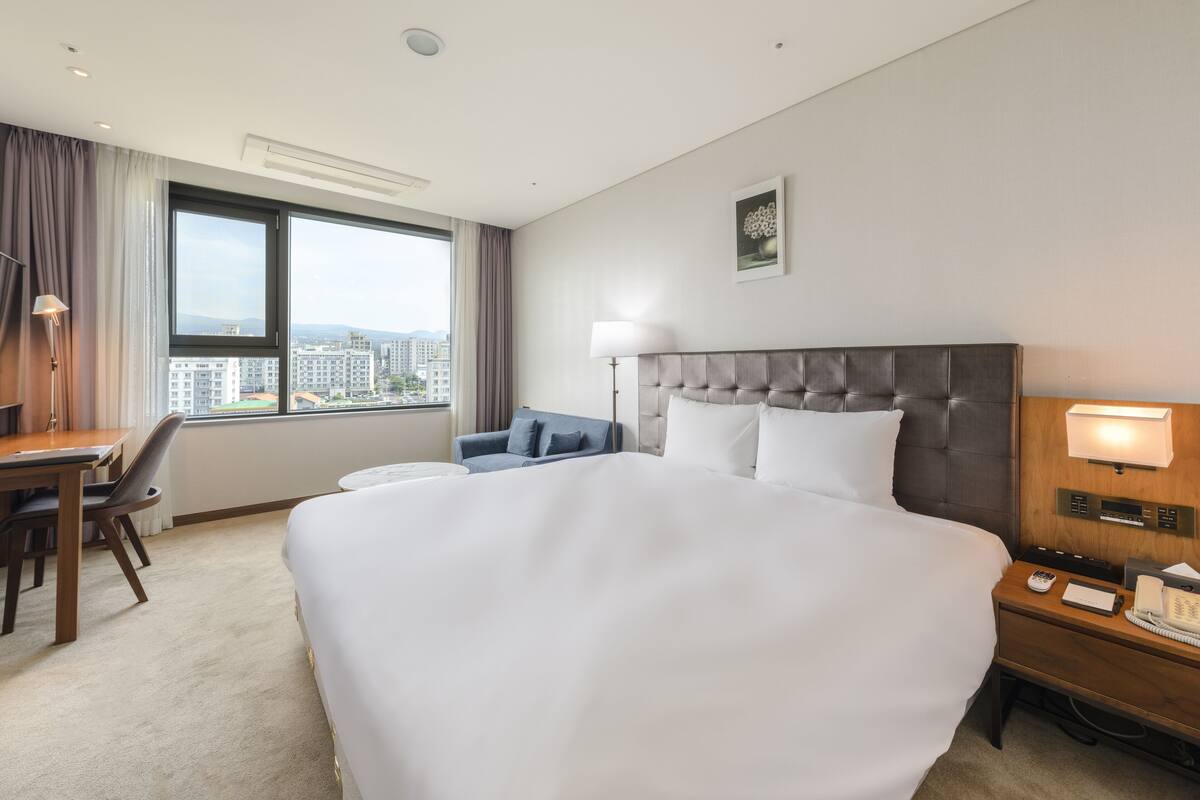 Hotelkamer met een groot bed, bureau, stoel, blauwe bank, nachtkastje met lamp en raam met uitzicht op de stad.