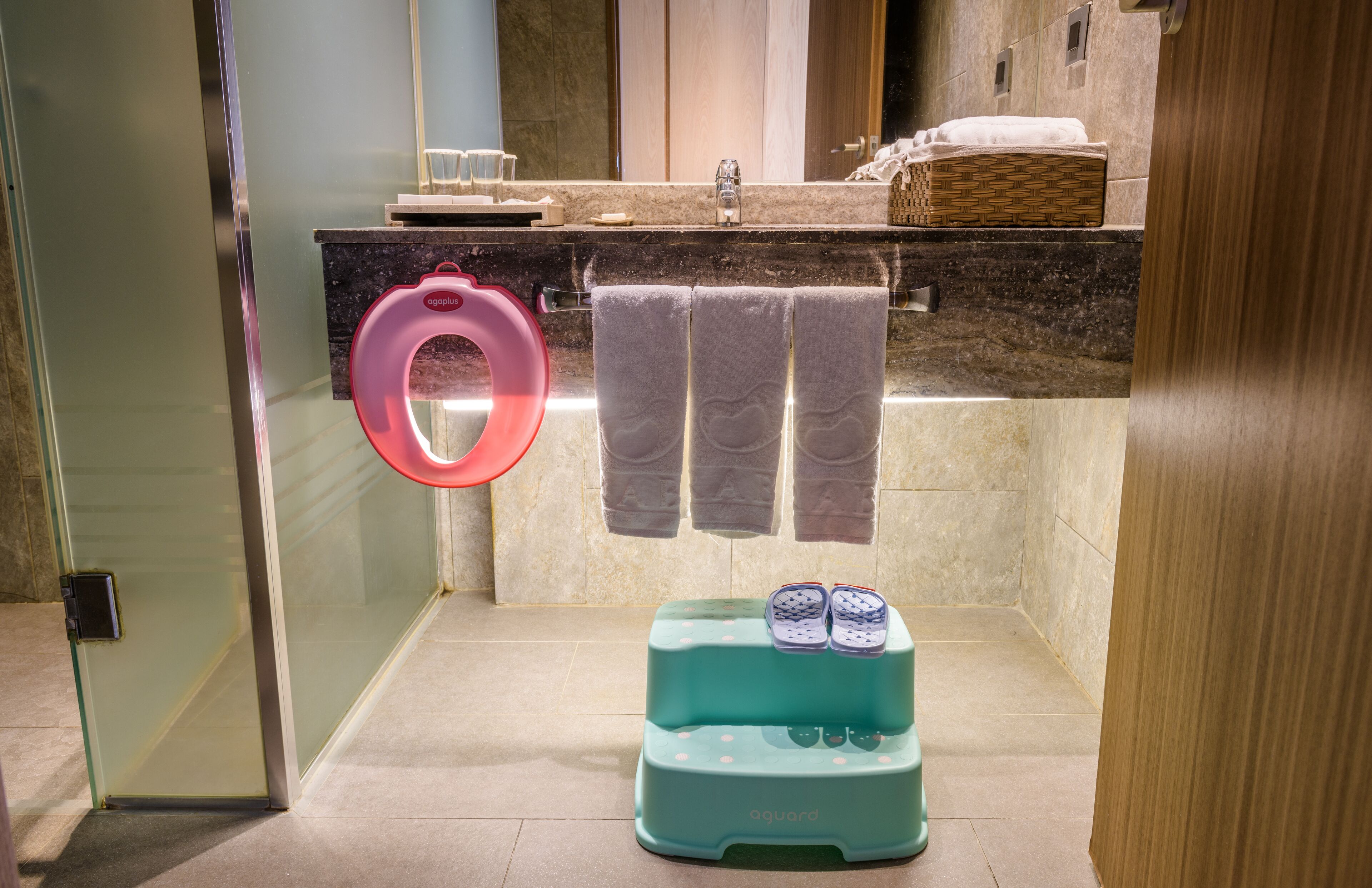 Un baño con cabina de ducha de vidrio, lavamanos con encimera de mármol y un taburete con pantuflas.