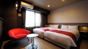 A hotel room with a bed, a red chair, a small table, an air conditioning unit, and a window with curtains.
