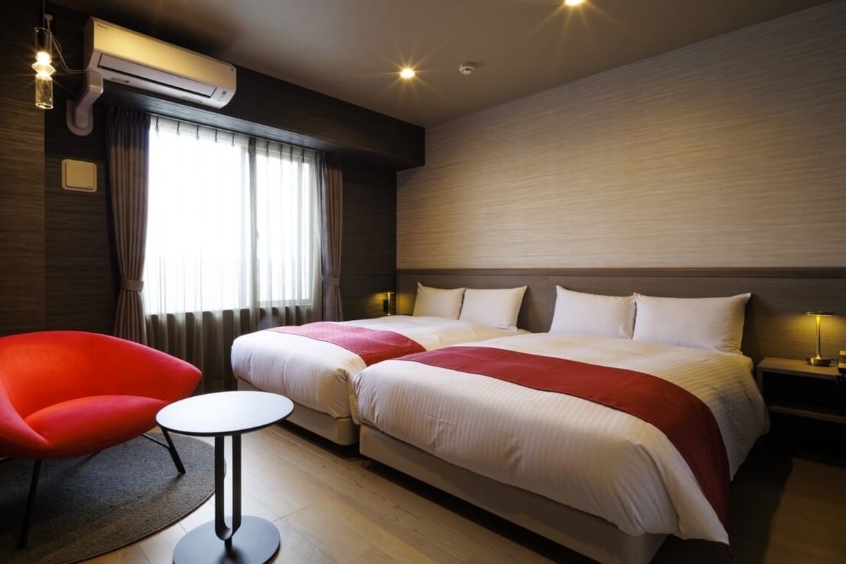 Habitación de hotel con dos camas, una silla roja, una mesita pequeña y un equipo de aire acondicionado.