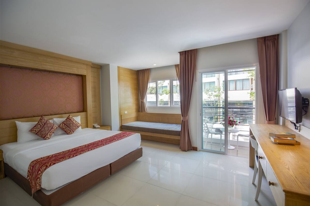 A hotel room with a large bed, a desk, a television, a window with curtains, and a balcony with a table and chairs.
