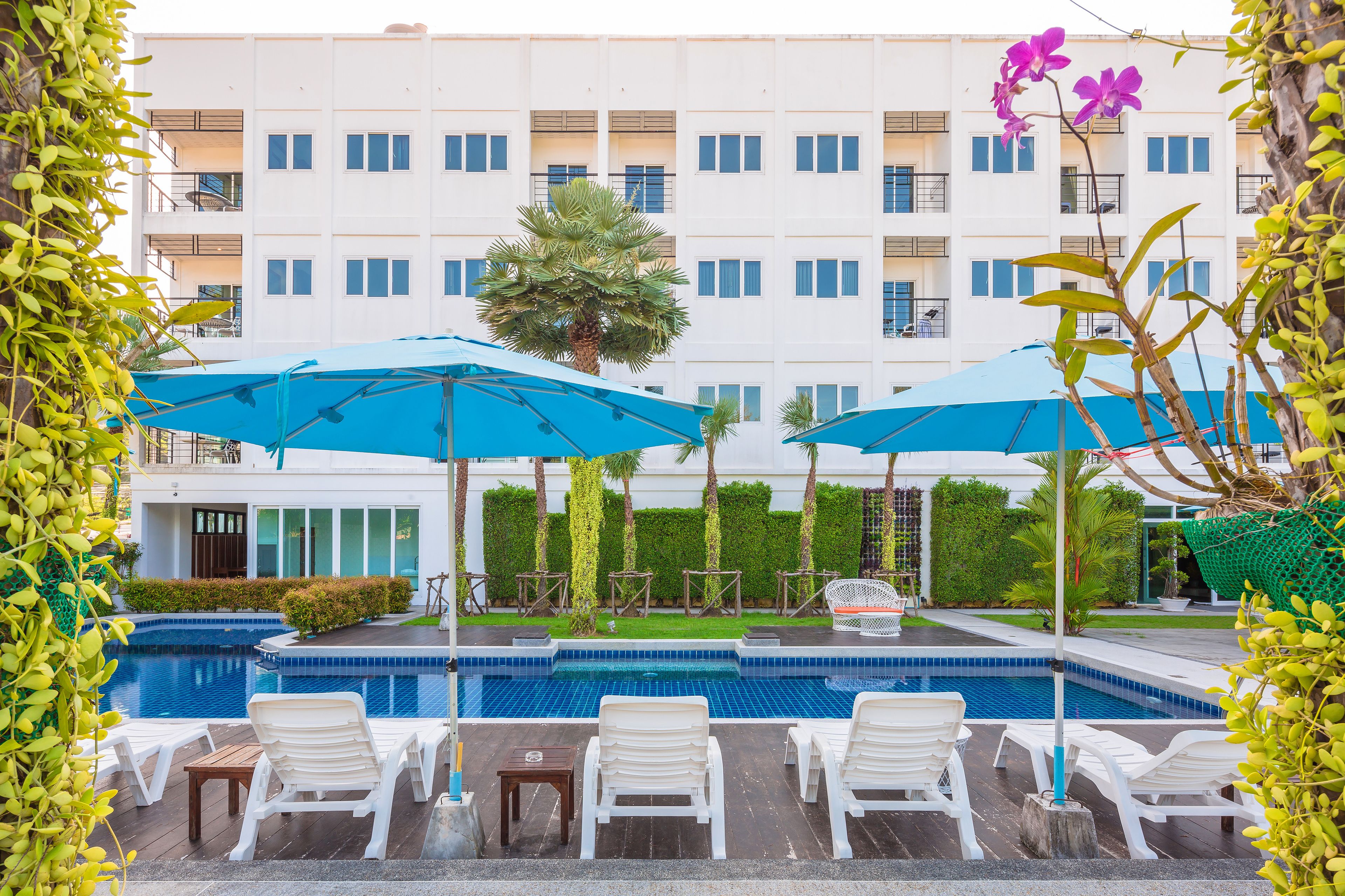 A pool area with lounge chairs and umbrellas, surrounded by a building with balconies and a well-maintained garden.