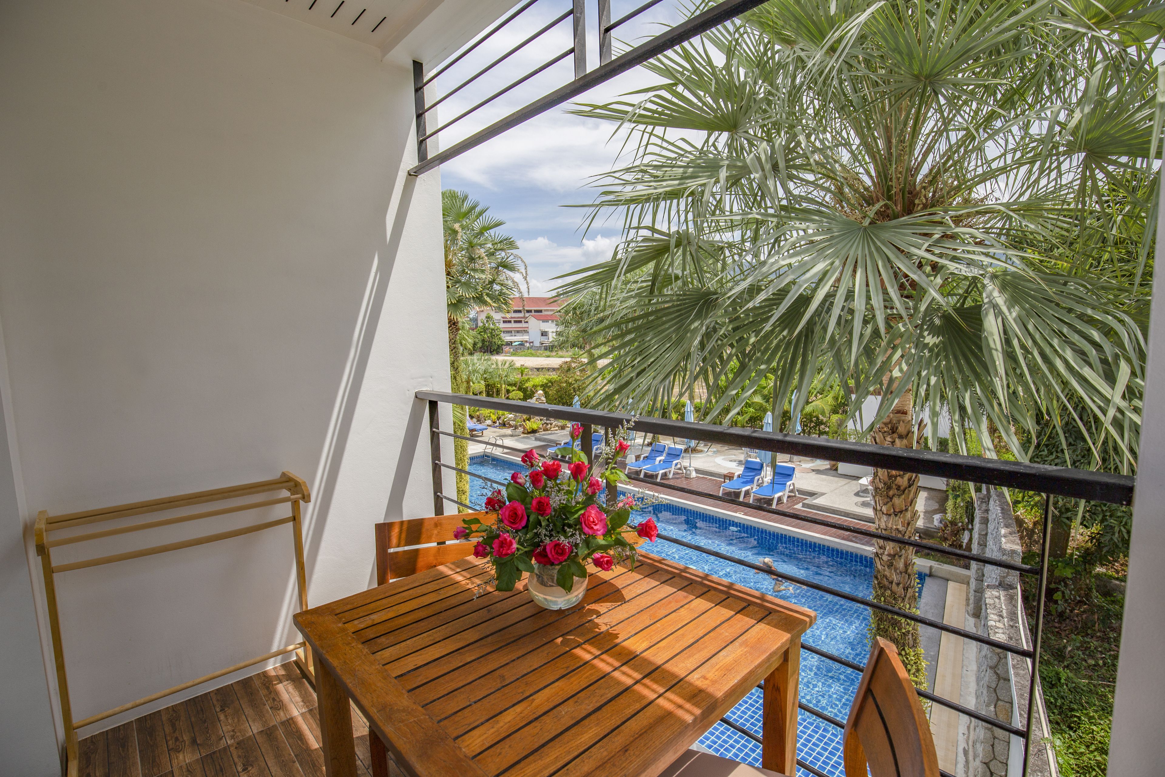 A balcony with a wooden table and chairs, a flower vase, and a view of a swimming pool and palm trees.