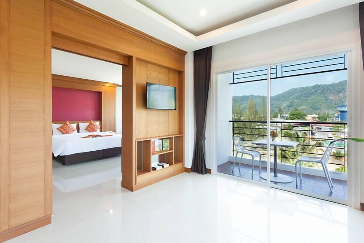 A modern hotel room with a large bed, a TV, a balcony with outdoor seating, and a view of the mountains.
