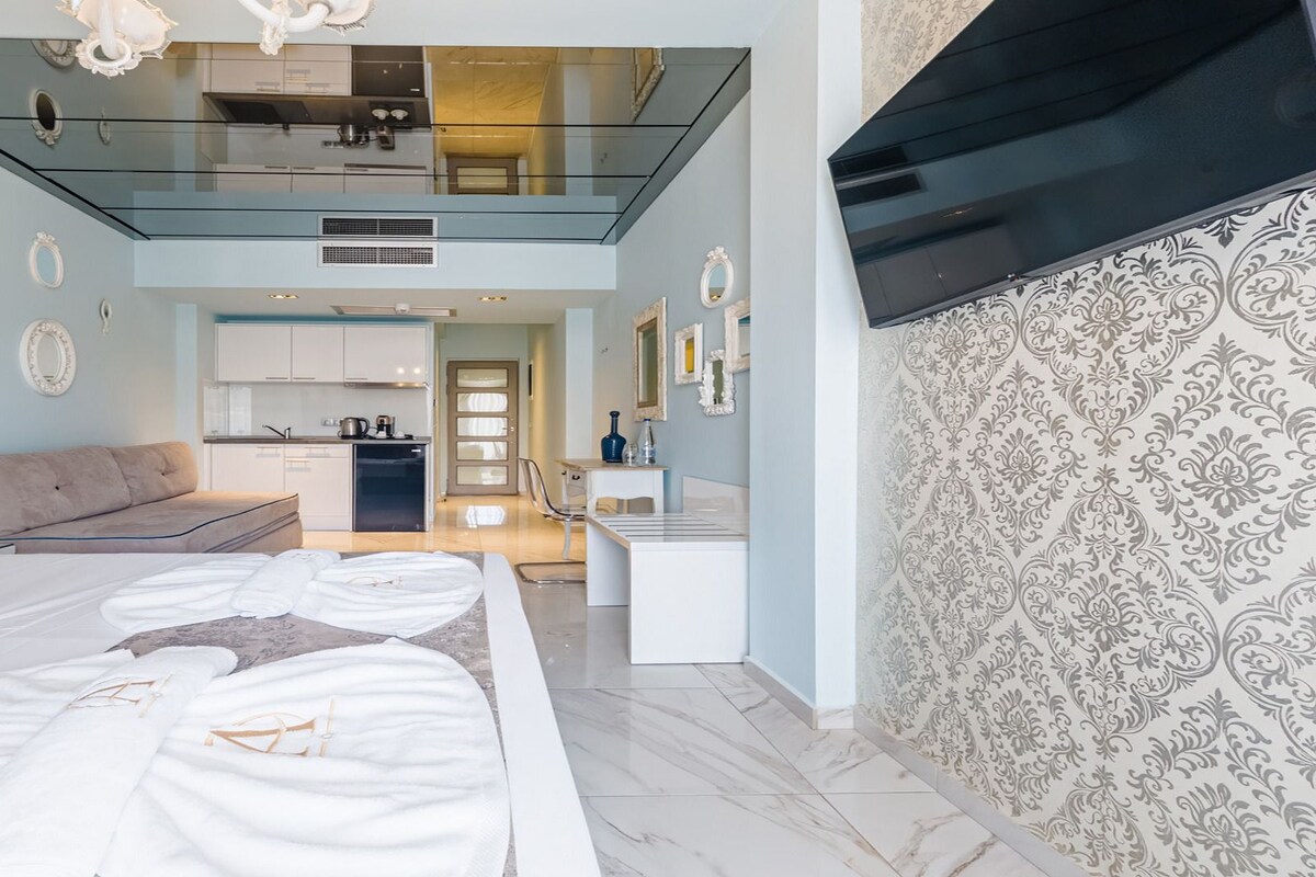 A modern bedroom with a large bed, a sofa, a kitchen area, and a TV.