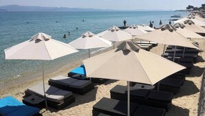 A beach with rows of sunbeds and umbrellas, clear blue water, and a distant shoreline. - Alpha Luxury Suites (Kassandra)