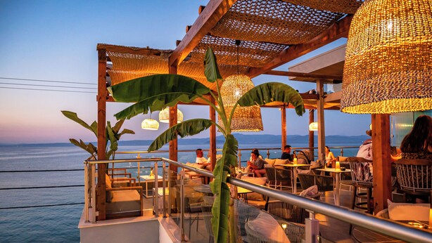 An outdoor dining area with tables and chairs, a railing, and a view of the ocean. - Alpha Luxury Suites (Kassandra)