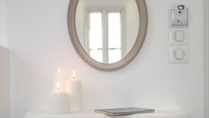 Un baño con lavamanos pedestal blanco, espejo redondo con marco floral, dos velas encendidas, un libro y un taburete estampado con motivos florales.