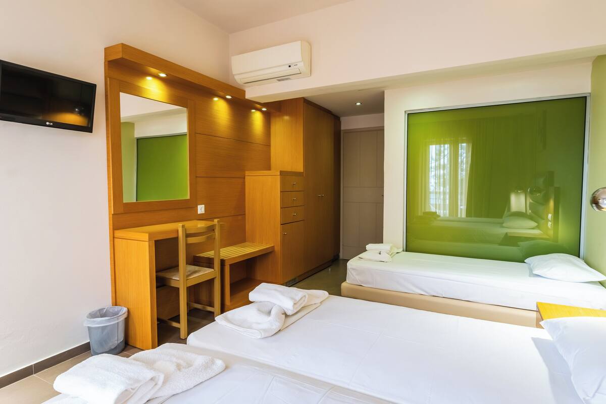 Habitación de hotel con dos camas, un escritorio, una silla, un espejo y una ventana con tonalidad verdosa.