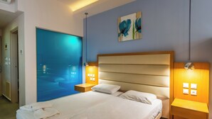 Un cuarto de hotel con una cama grande, dos mesitas de noche, una obra de arte en la pared y vista al mar a través de un ventanal.