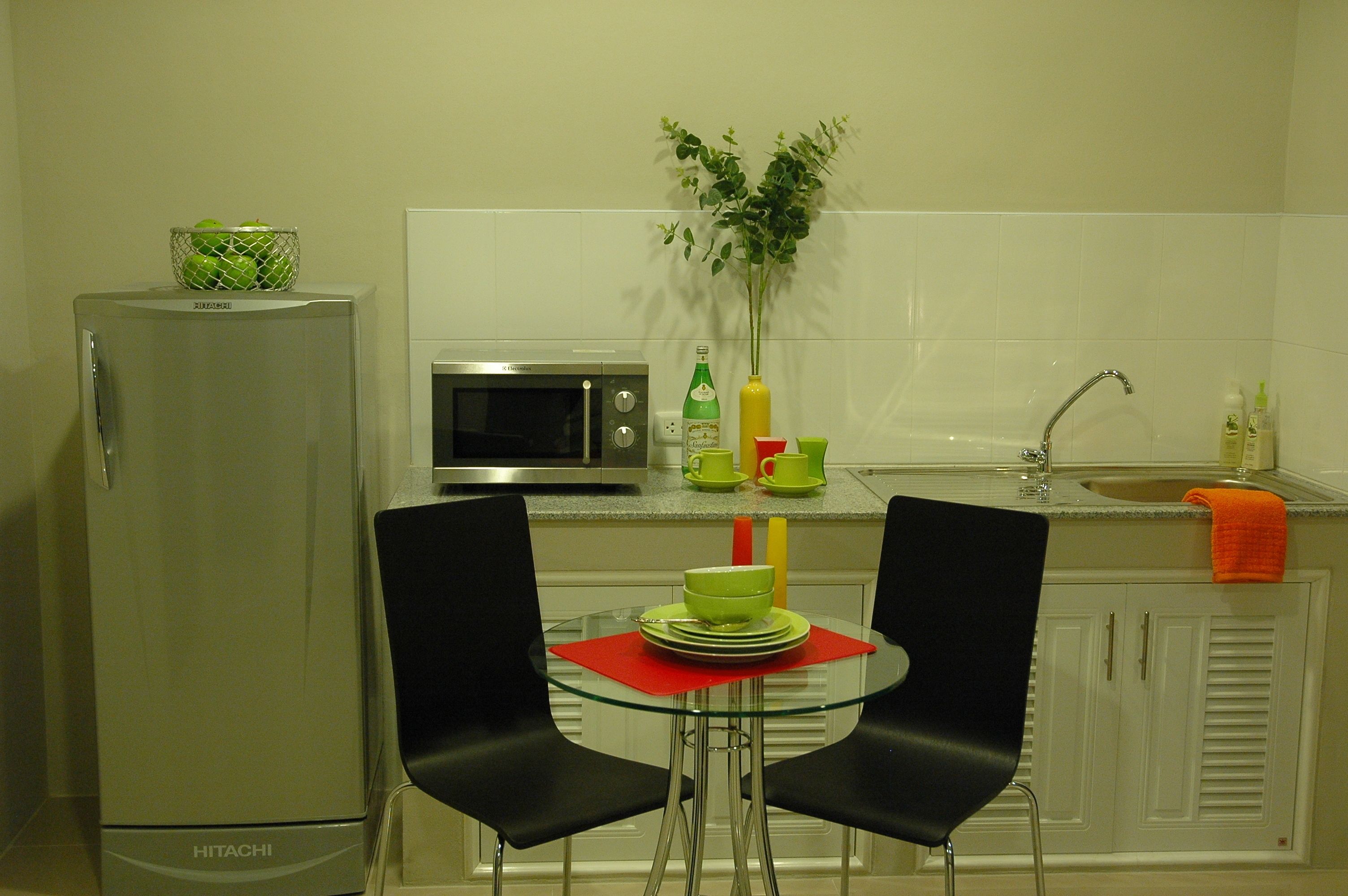 A compact kitchen with a refrigerator, microwave, and sink. There are two black chairs around a small glass table with a green bowl and plates.