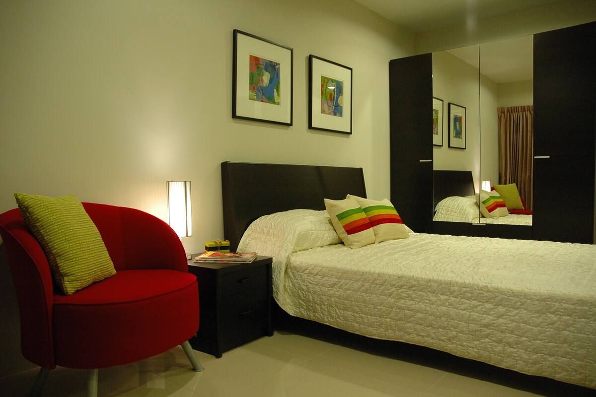 A hotel room with a bed, a red armchair, a nightstand, and two framed artworks on the wall.