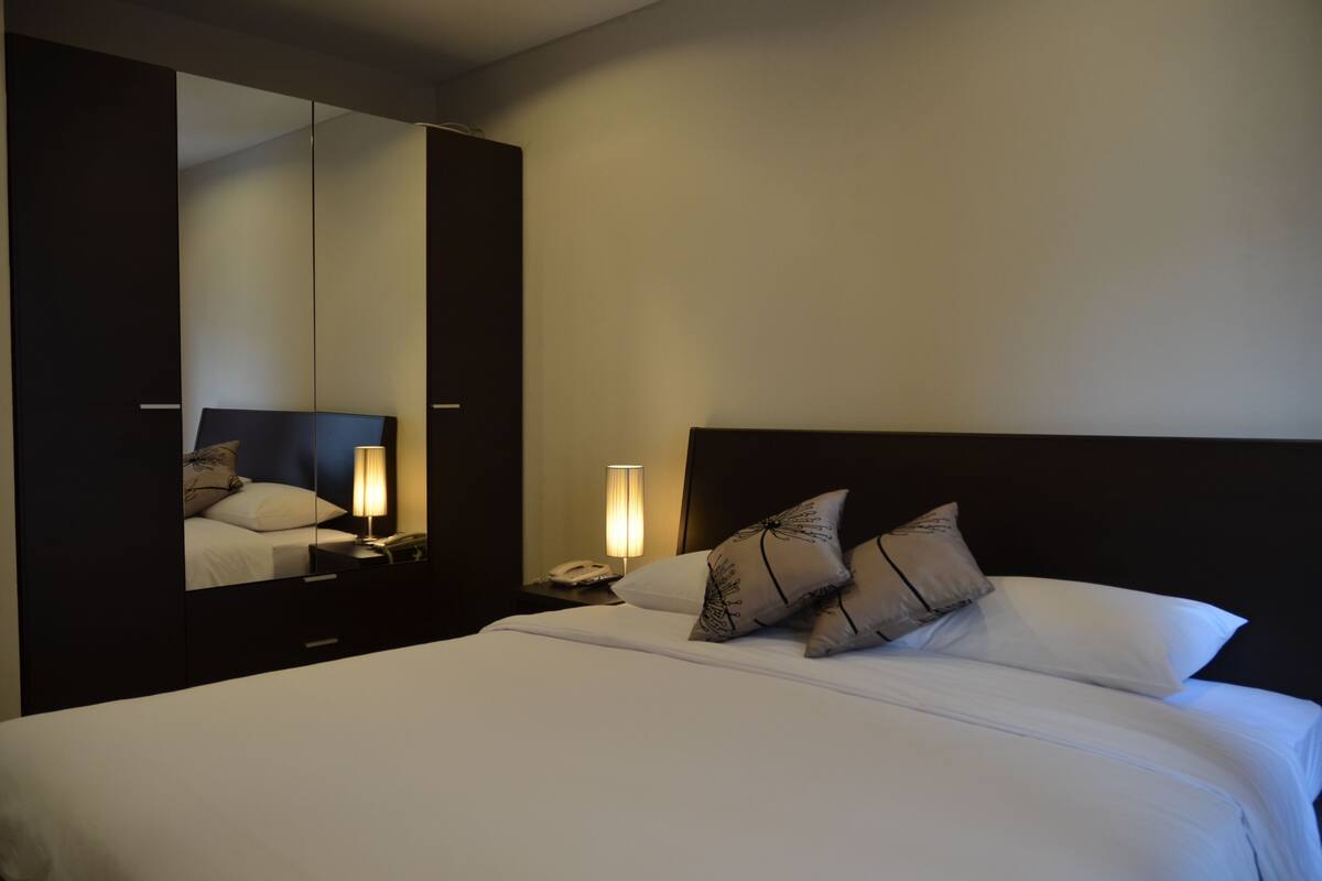 A hotel room with a large bed, a nightstand with a lamp, a mirror, and a wardrobe.
