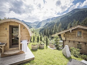 Una cabaña de madera con entrada circular y terraza, rodeada de un paisaje montañoso y vegetación.