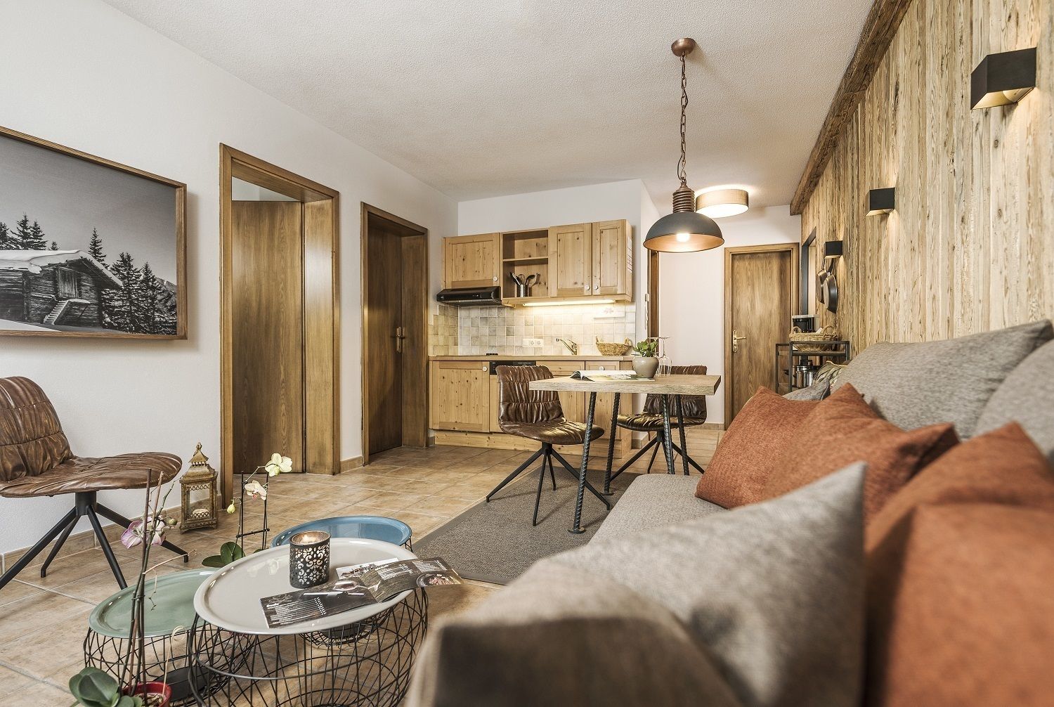 A cozy living room with a sofa, a coffee table, and a dining area with a table and chairs. There is a kitchen with wooden cabinets and a hanging light fixture.