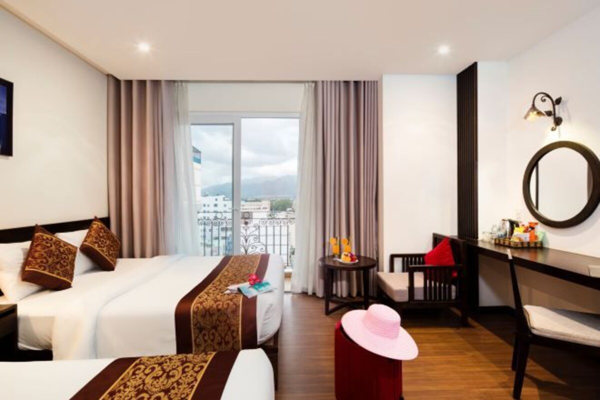 A hotel room with a large bed, a desk, a chair, a small table with a lamp, and a view of the city through the window.
