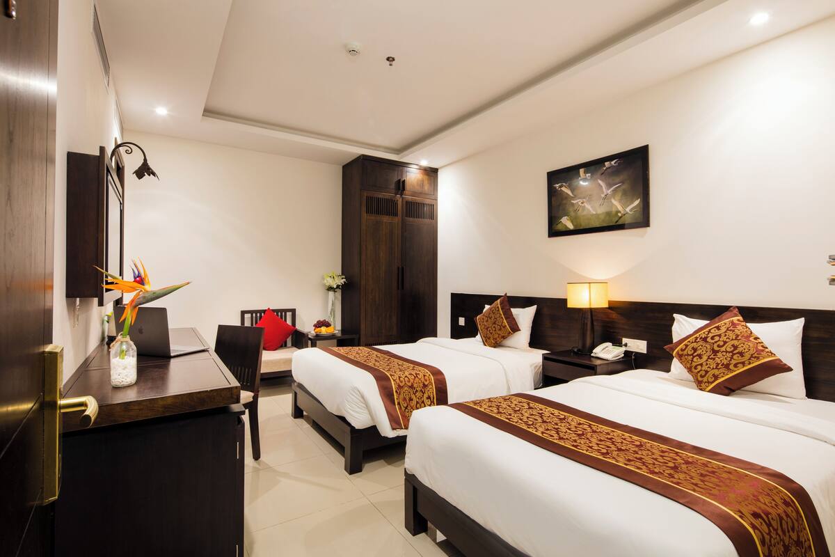Quarto de hotel com duas camas, uma escrivaninha, uma cadeira, um guarda-roupa, mesa de cabeceira com abajur e um quadro na parede.