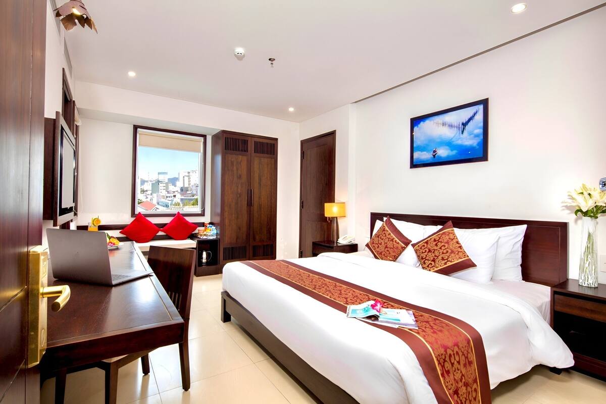 Quarto de hotel com cama, escrivaninha com laptop, janela com vista para a cidade e um quadro na parede.