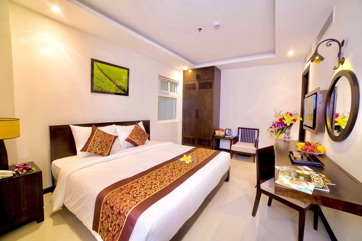Quarto de hotel com uma cama grande, uma escrivaninha com cadeira, um espelho e uma janela.