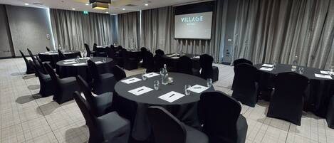 A conference room with multiple round tables, black chairs, and a projector screen displaying "VILLAGE HOTELS".