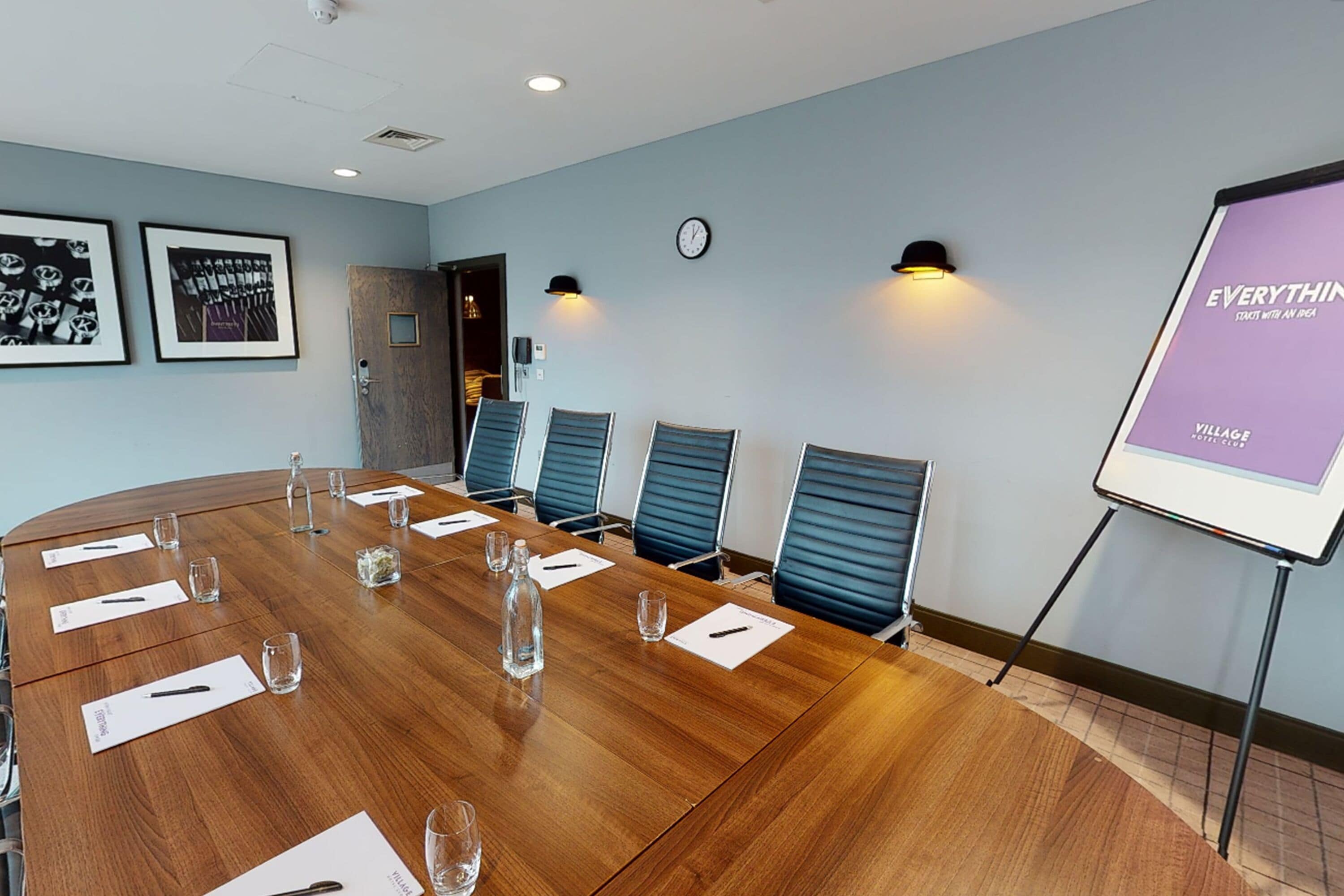A conference room with a wooden table, blue chairs, a projector screen, and framed pictures on the wall.