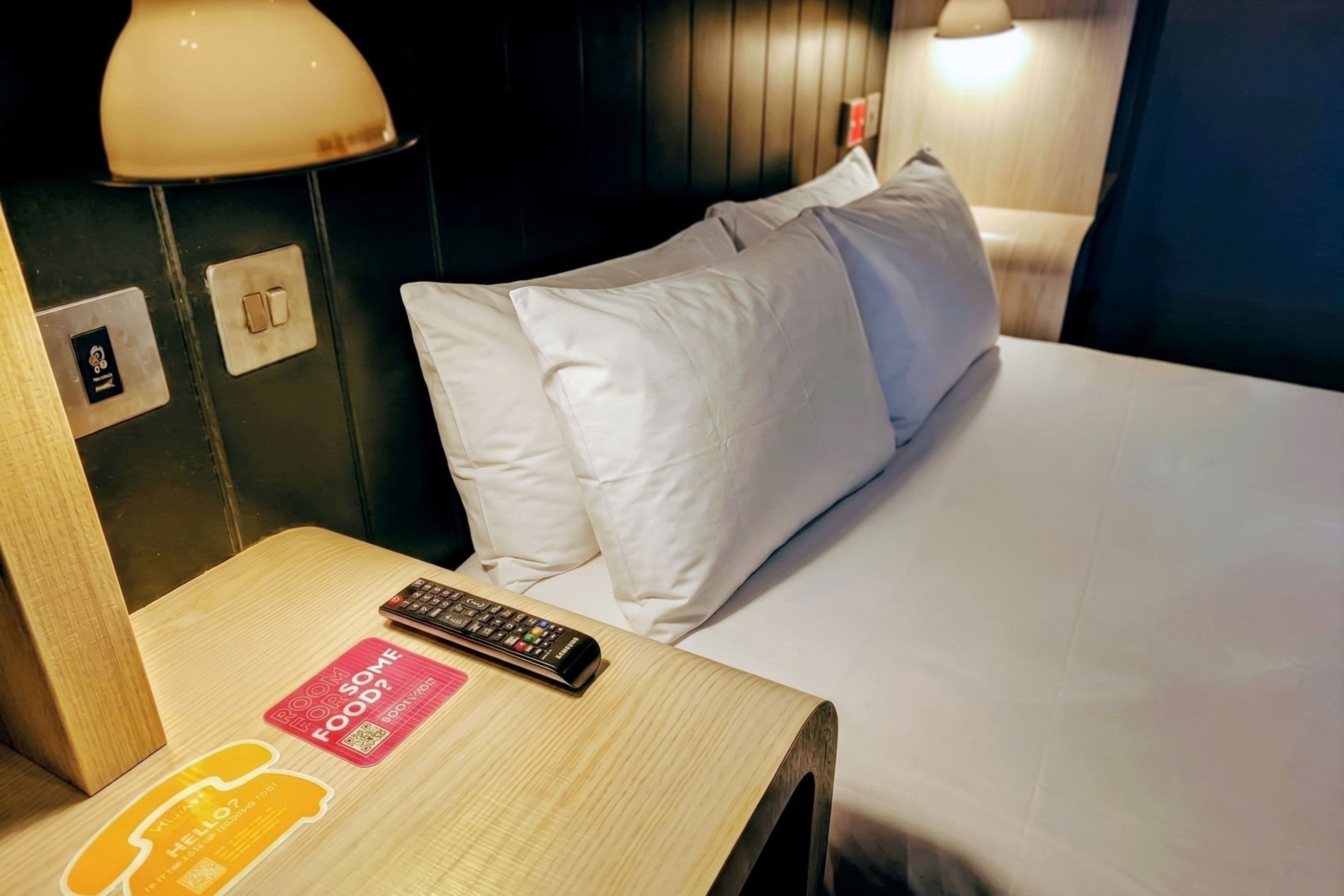 Habitación de hotel con cama, almohadas, mesita de noche, control remoto y lámpara fijada a la pared.