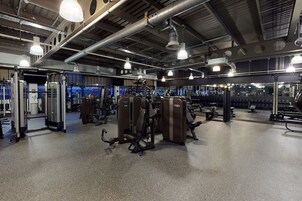 A gym with various exercise machines, large windows, and industrial-style lighting.