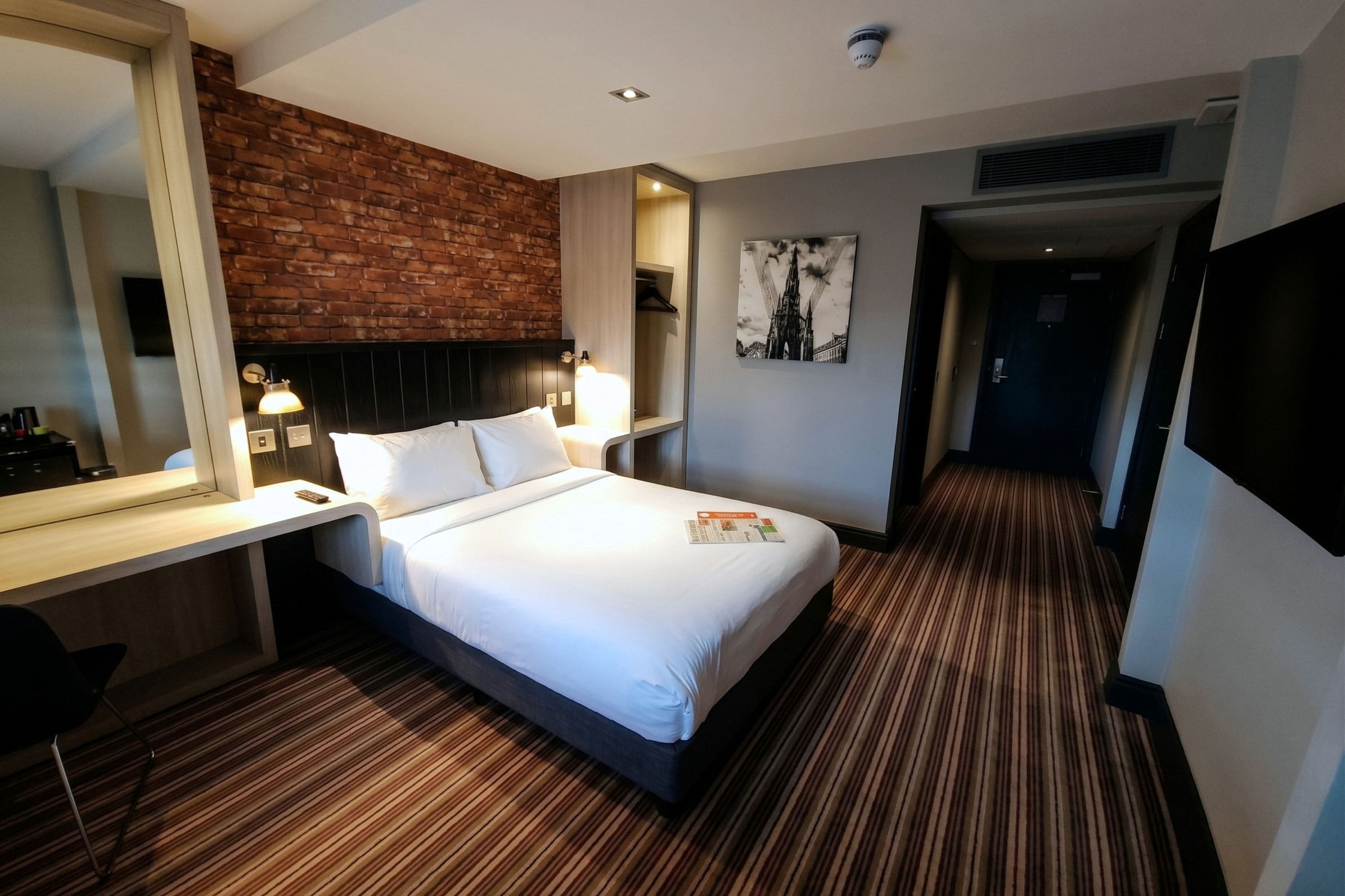Een hotelkamer met een bed, een bureau, een spiegel, een aan de muur hangend schilderij en een deur naar een andere kamer.