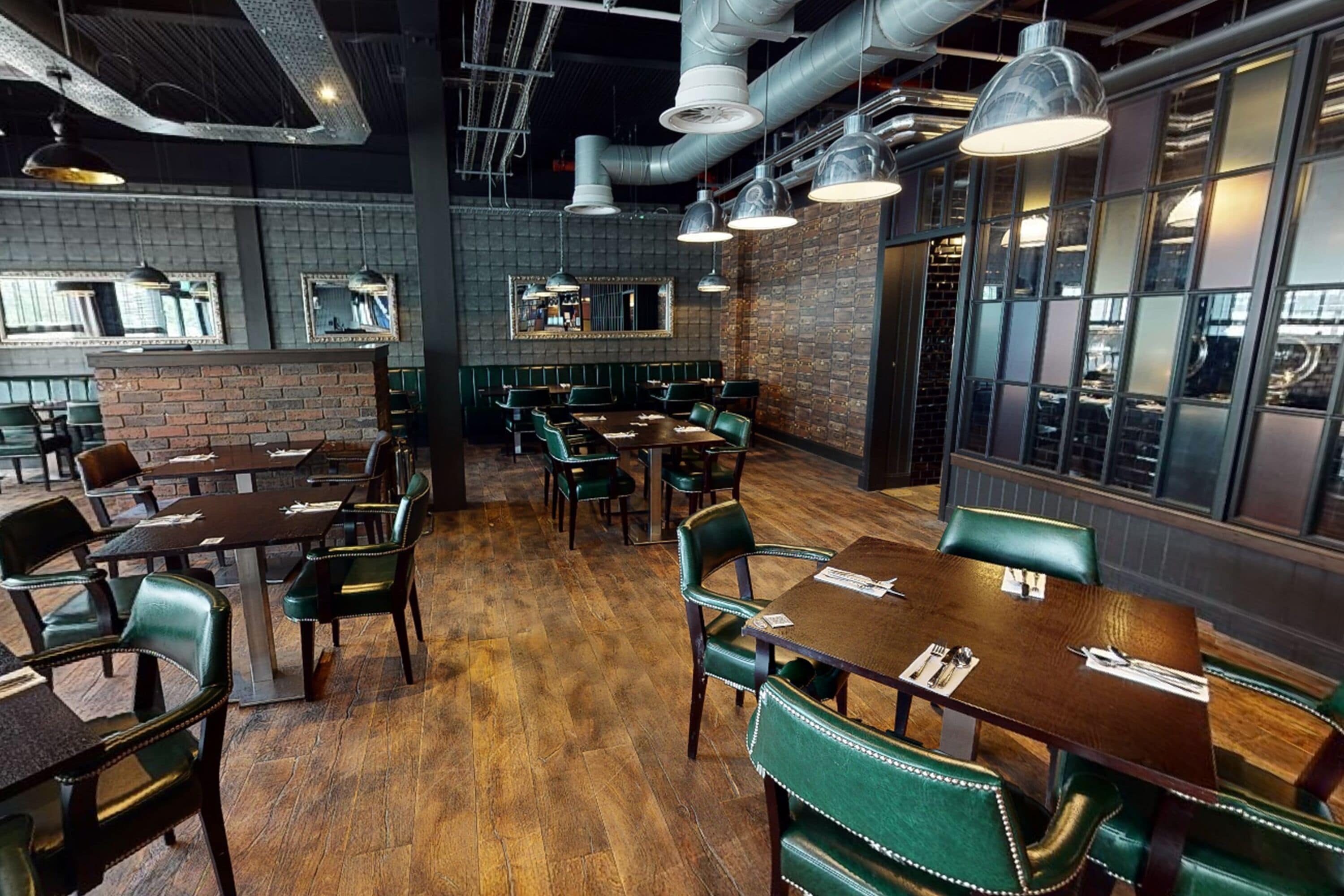 Een restaurantinterieur met bakstenen muren, houten vloeren en groene bekleden stoelen.
