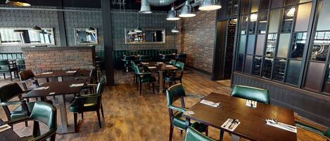 Een restaurantinterieur met bakstenen muren, houten vloeren en groene bekleden stoelen.