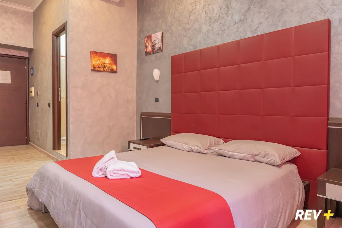 Habitación de hotel con una cama grande, cabecera roja, suelo de madera, una puerta y una lámpara fijada a la pared.