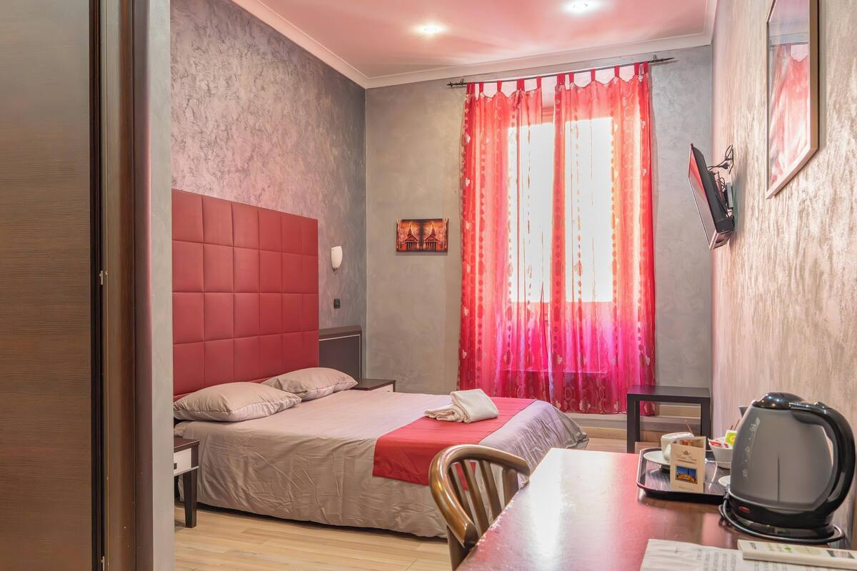 Habitación de hotel con cama, escritorio con hervidor, silla, televisor y ventana con cortinas rojas.