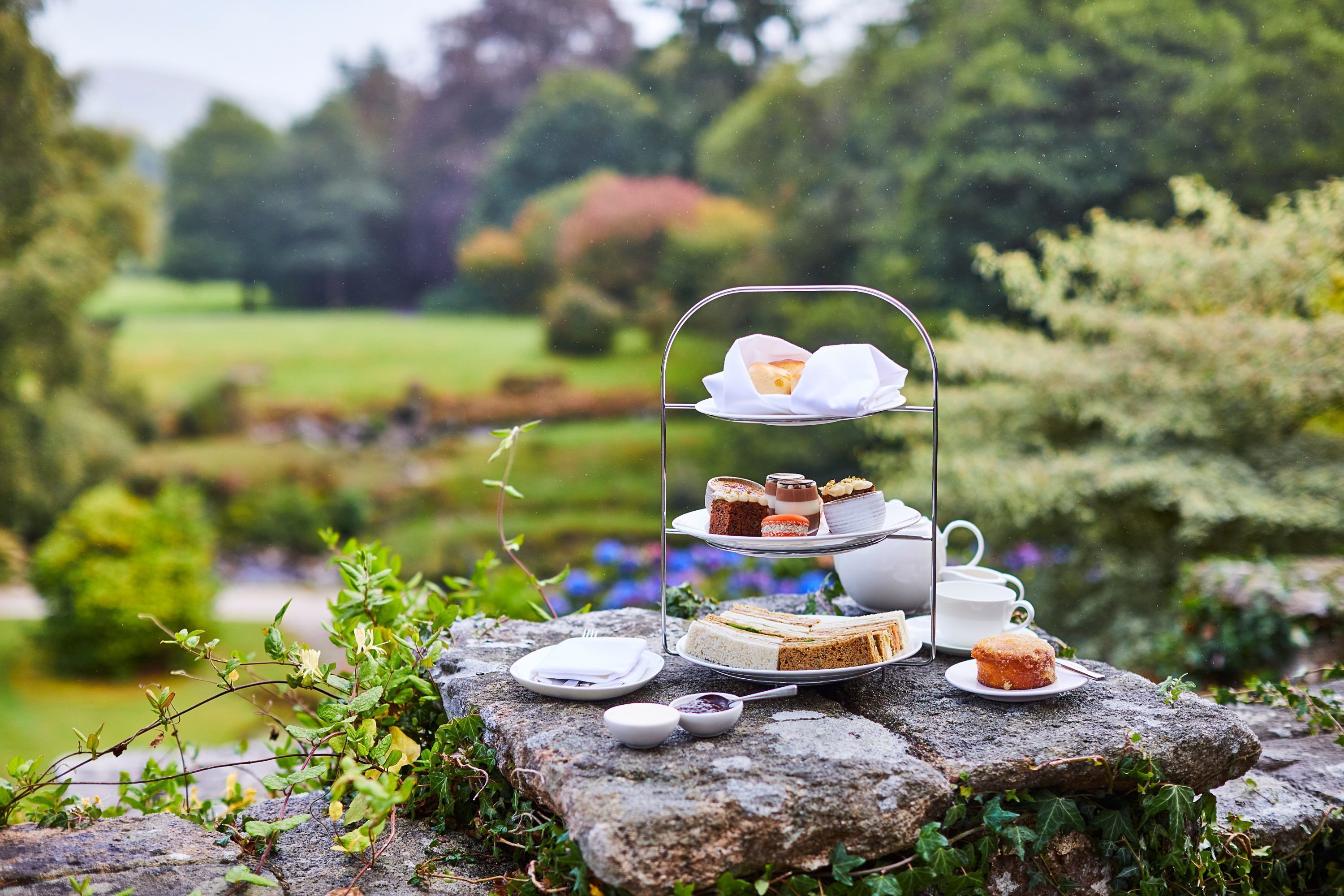 A three-tiered outdoor tea service on a stone table surrounded by greenery.