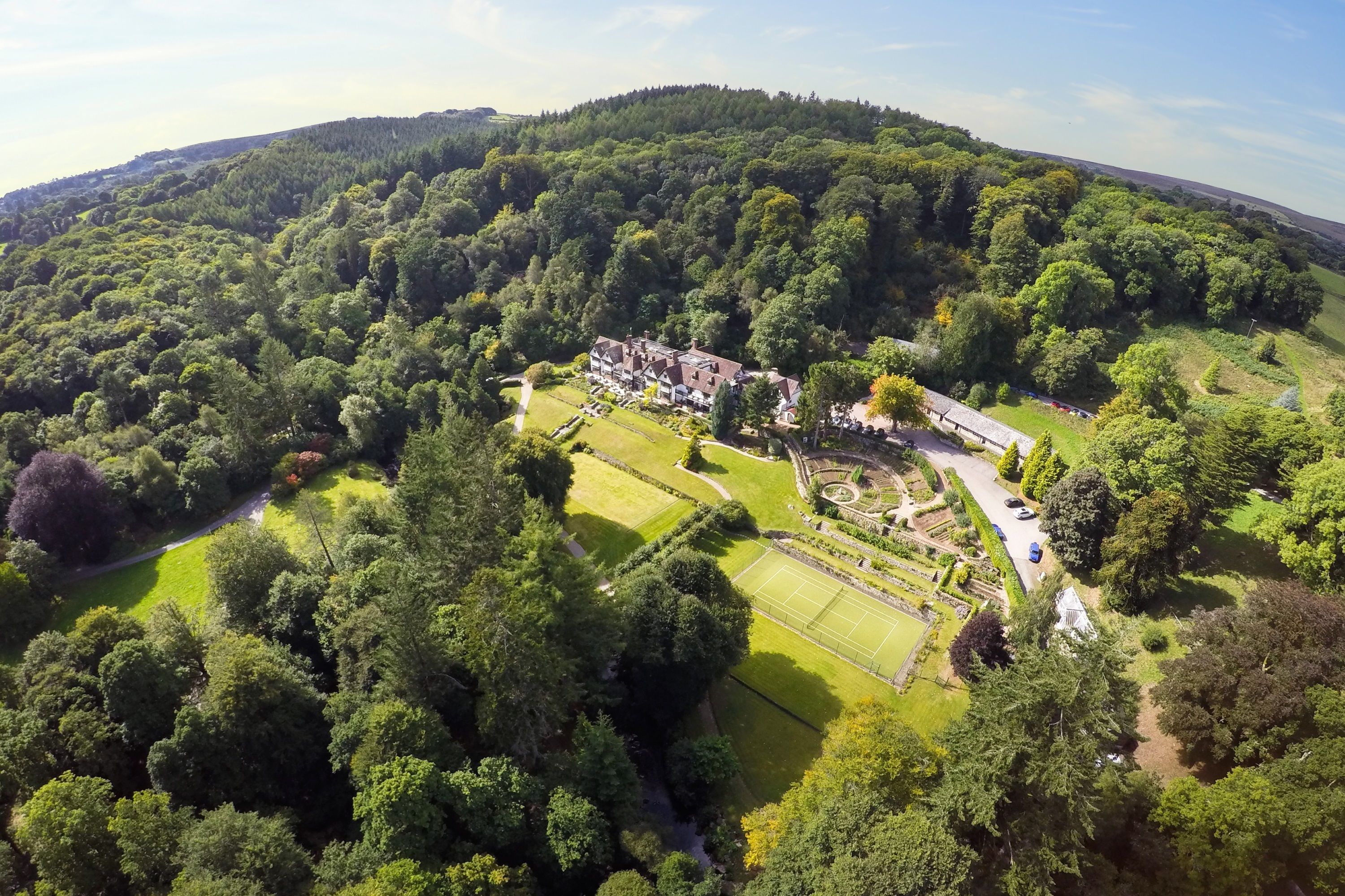 An aerial view of a large estate with a tennis court, landscaped gardens, and a variety of trees.