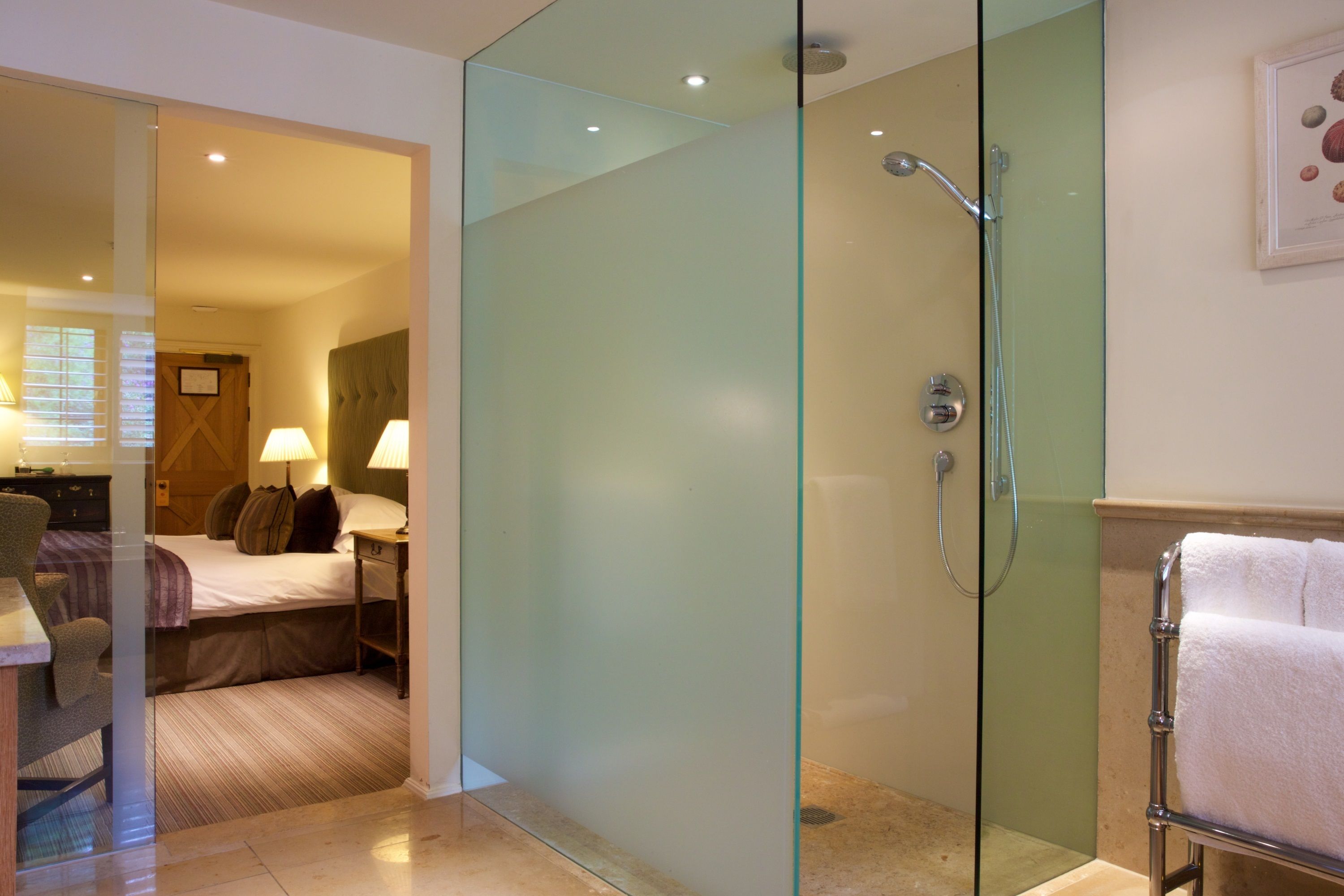 Habitación de hotel con ducha de vidrio, cama con almohadas, mesita de noche con lámpara y perchero.
