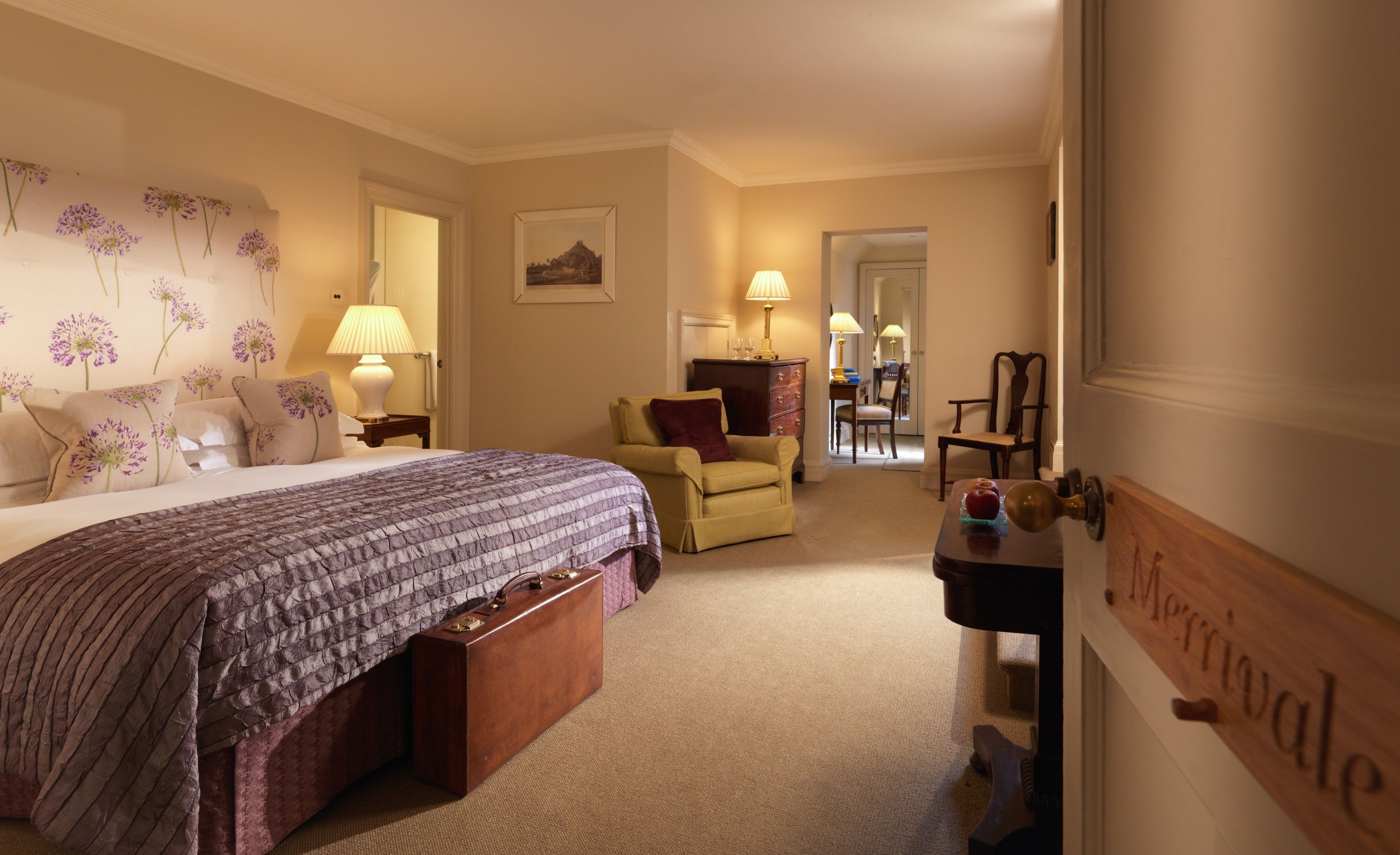 A hotel room with a large bed, a chair, a small table with a lamp, and a wooden door with a nameplate.