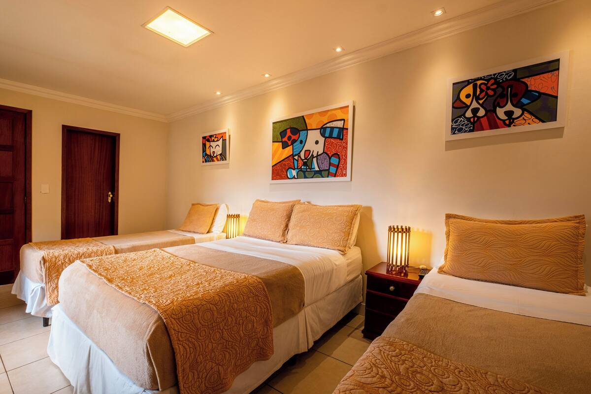 A hotel room with three beds, each with a patterned bedspread, a nightstand with a lamp, and artwork on the walls.
