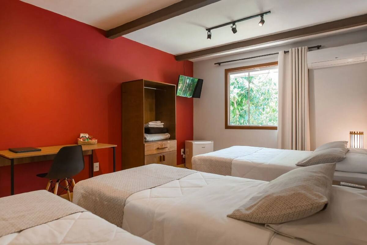 A hotel room with a red wall, a desk with a computer, two beds, a TV, and a window with curtains.