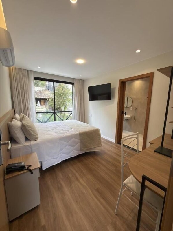 A modern hotel room with a bed, a desk, a chair, a TV, and a bathroom with a shower.