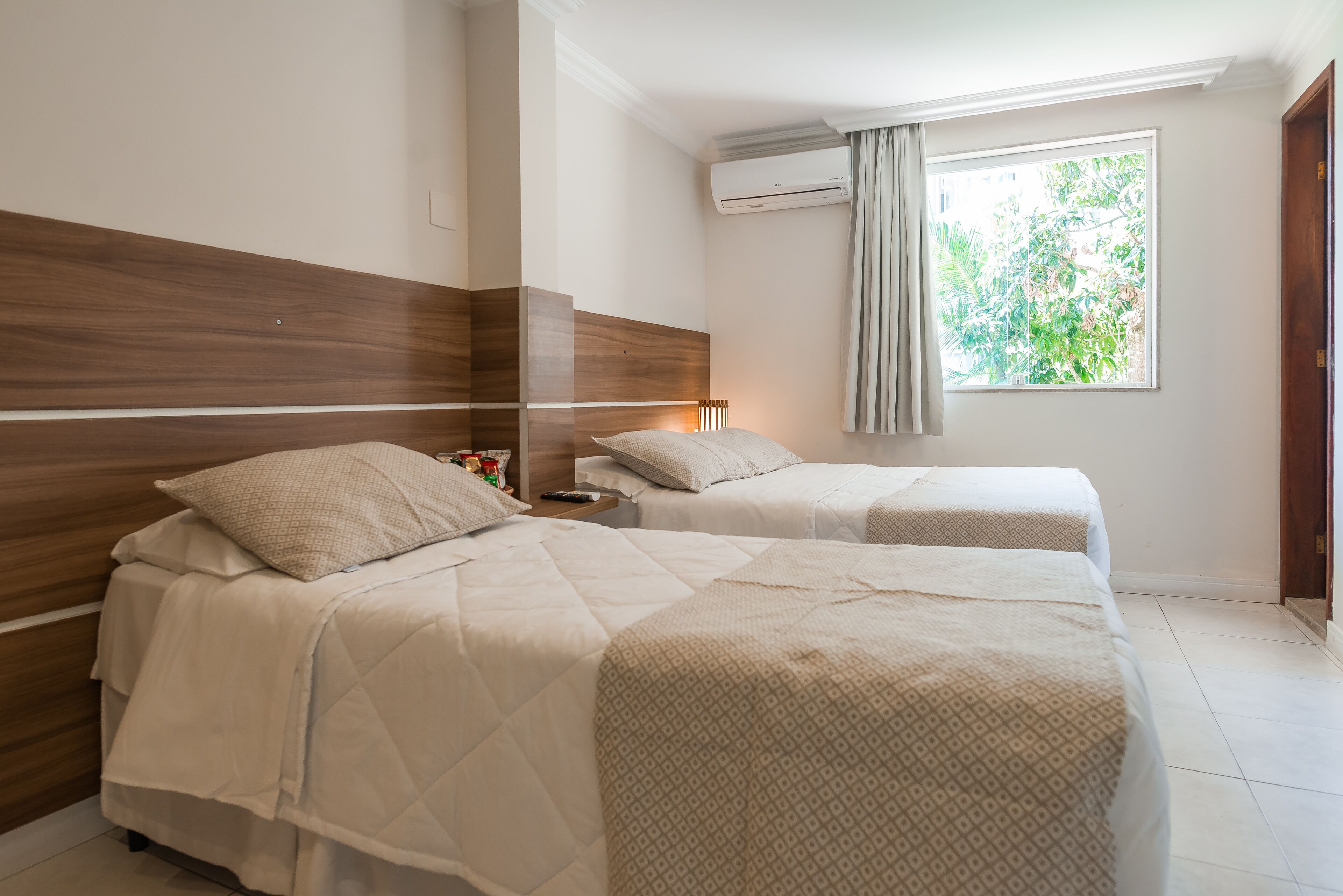 A hotel room with two beds, a wooden headboard, an air conditioning unit, and a window with curtains.