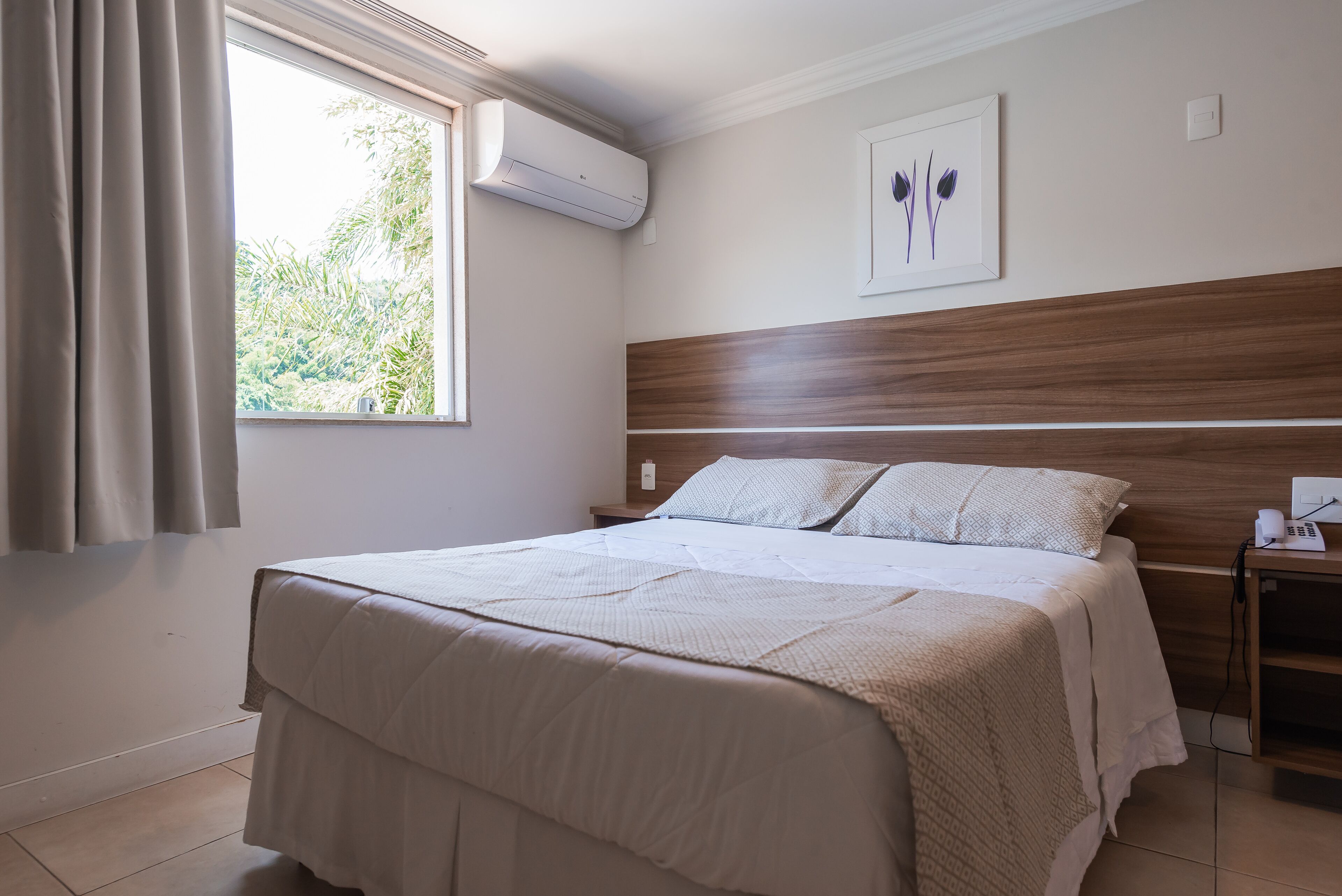 A neatly arranged bedroom with a bed, a nightstand, a wall-mounted air conditioner, and a window with curtains.
