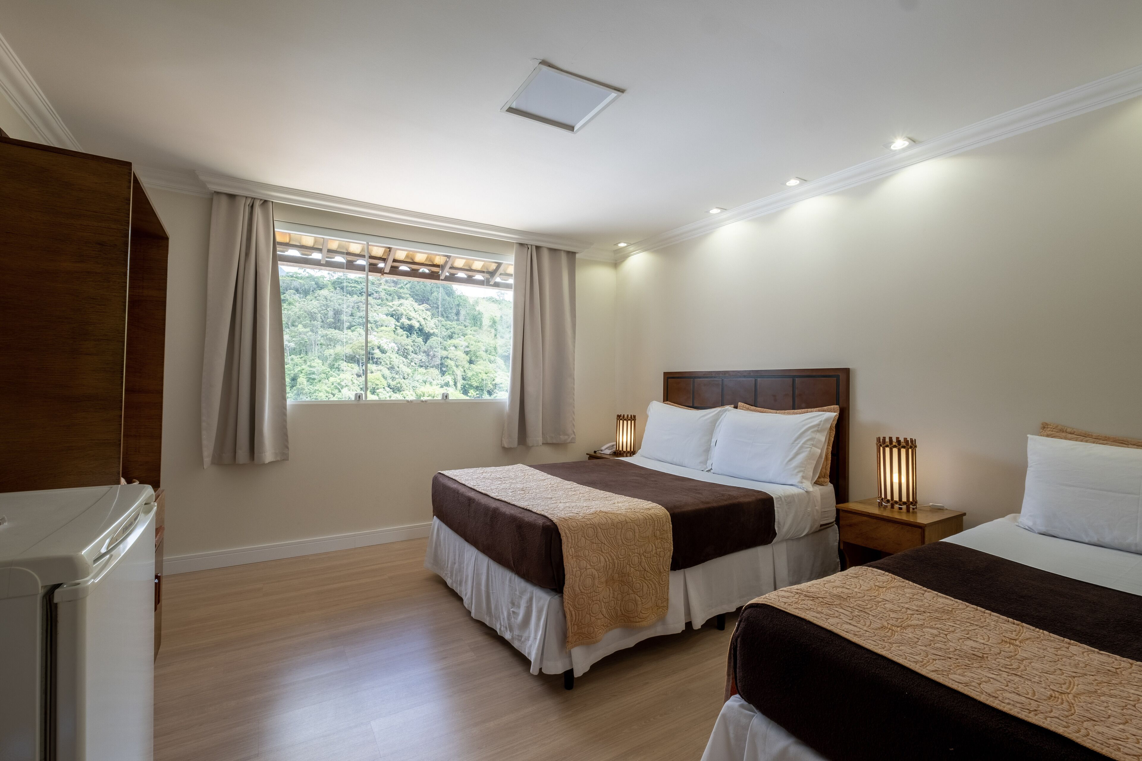 A bedroom with two beds, a wooden wardrobe, a window with curtains, and a view of greenery outside.